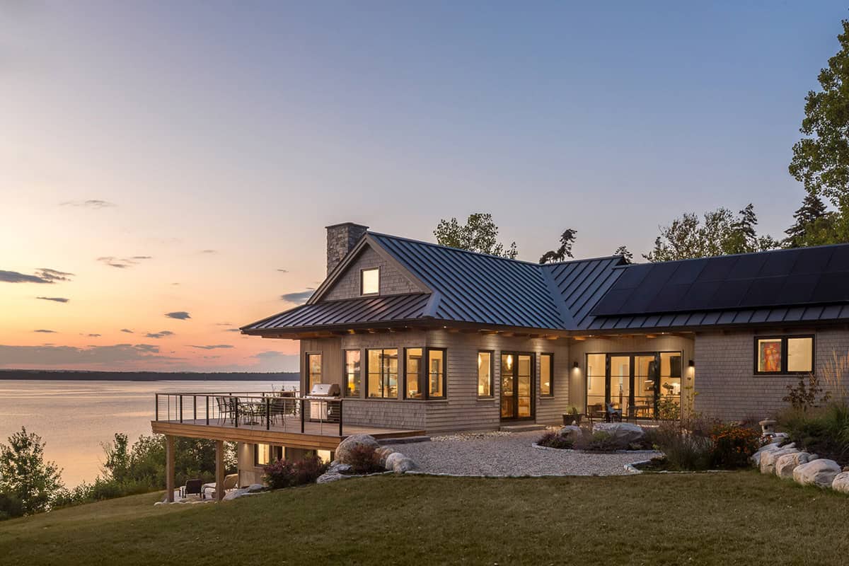 Shingled house with a dark metal roof and solar panels glowing at dusk beside a cable-railed deck overlooking the bay