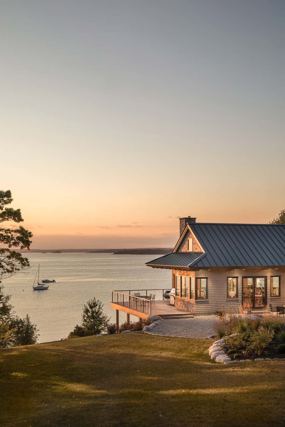 Lit-up house with a metal roof and deck at the water’s edge at sunset, with sailboats on the calm bay beyond