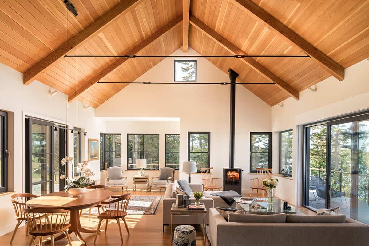 Open living and dining area under a vaulted wood ceiling with exposed beams, a black wood stove, and glass doors to the deck