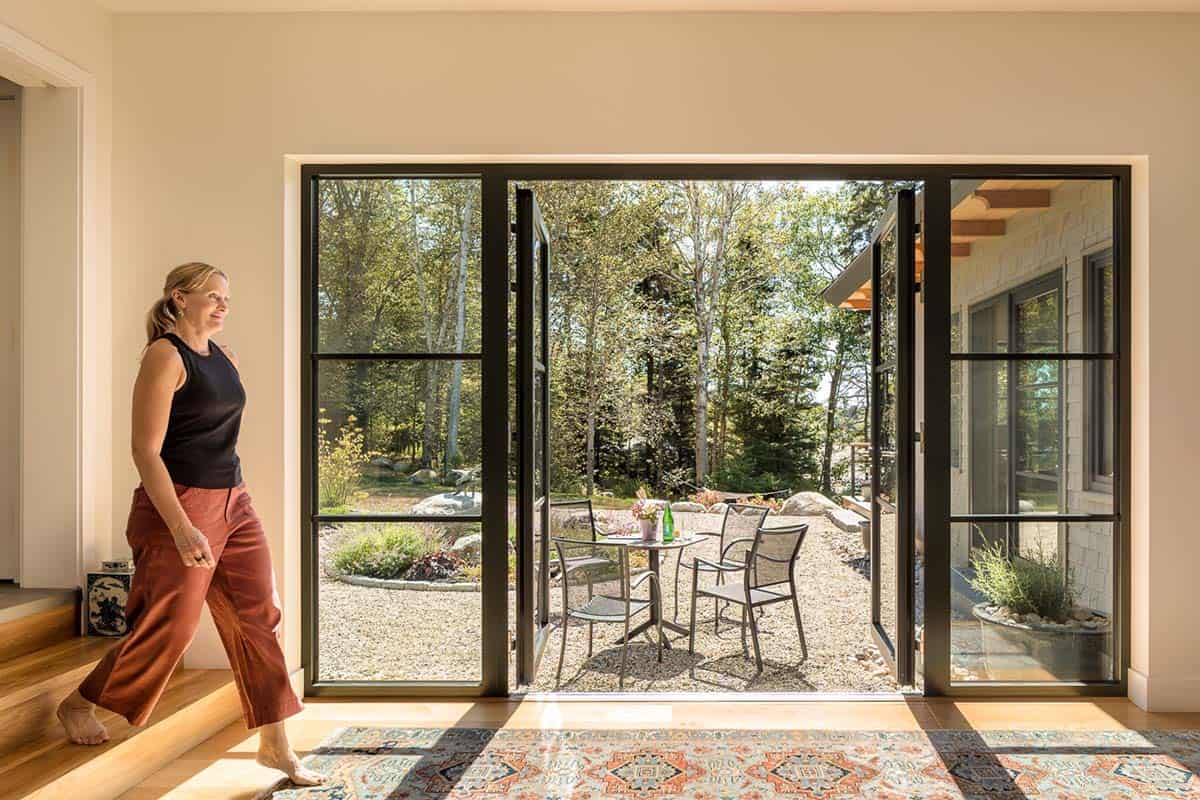 Woman walking past open steel-framed glass doors toward a gravel patio with a small table and chairs
