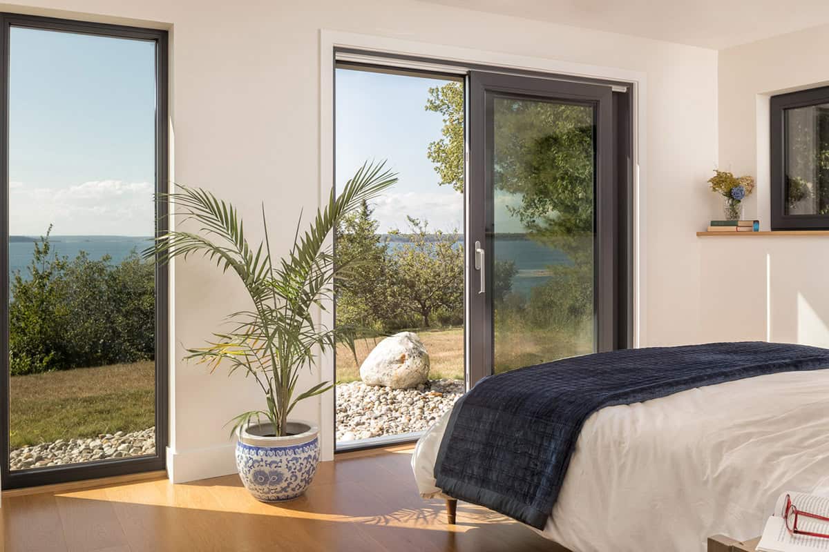Bedroom with a potted palm and sliding glass doors looking out to a boulder, lawn, and the water