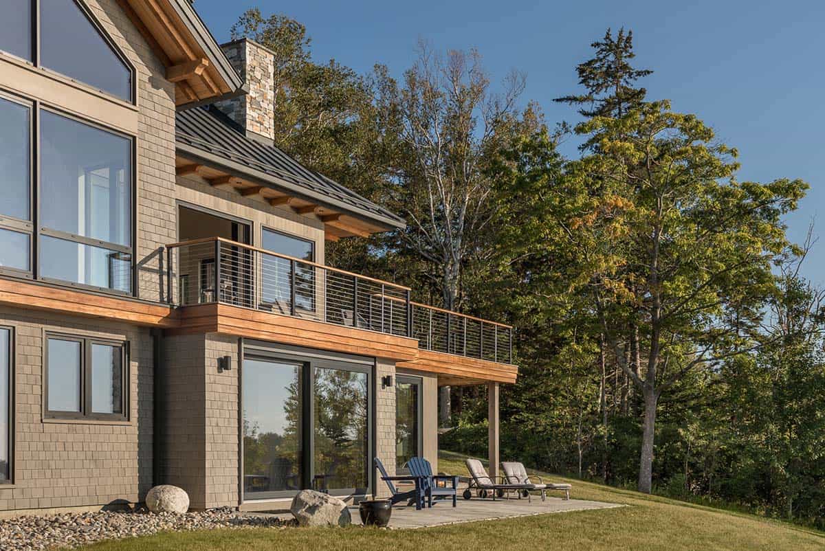Shingled house with a wood-trimmed upper deck and a ground-level patio with blue Adirondack chairs and lounge chairs
