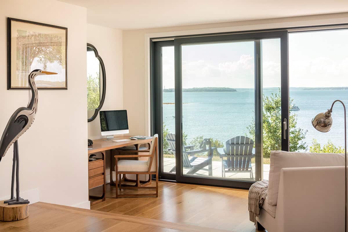 Home office nook with a wooden desk and a heron sculpture beside a sliding glass door overlooking Adirondack chairs and the bay