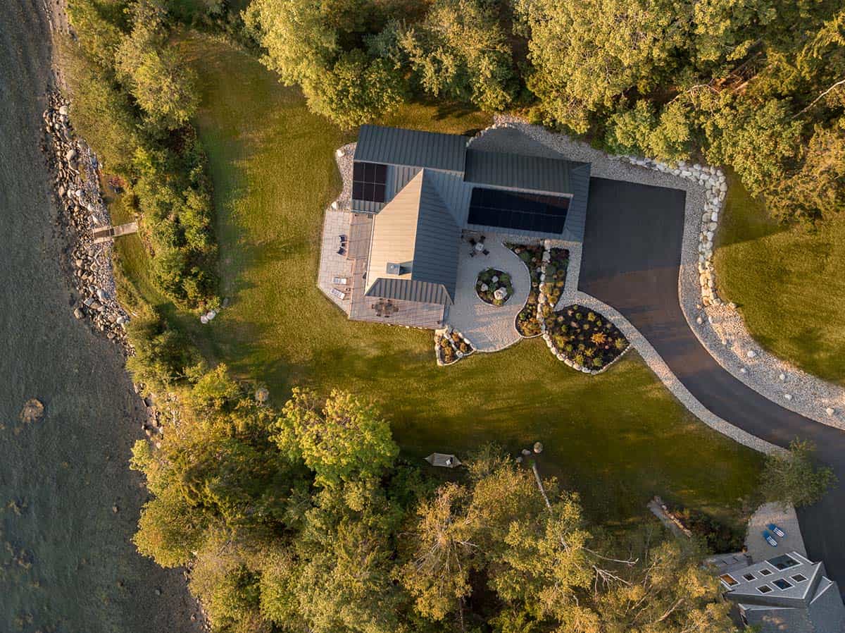 Aerial view of the house’s metal roof with solar panels, surrounded by lawn, gravel garden beds, a curving driveway, and the shoreline