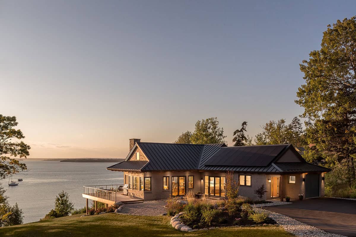 House with a deck and lit windows at dusk, with solar panels on the roof, overlooking the bay