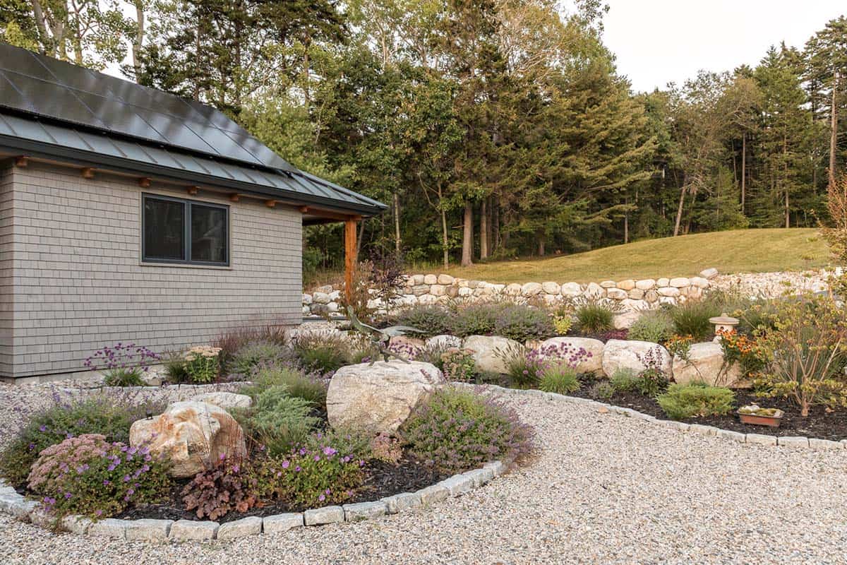 Garden beds with boulders and flowering perennials in a gravel courtyard beside the house, with a stone retaining wall behind