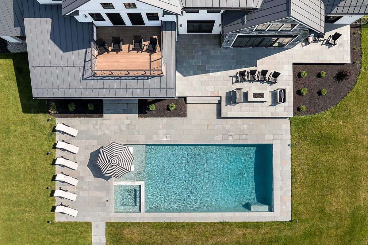 Overhead view of a rectangular pool with spa, white chaise lounges, striped umbrella, and bluestone patio with fire pit