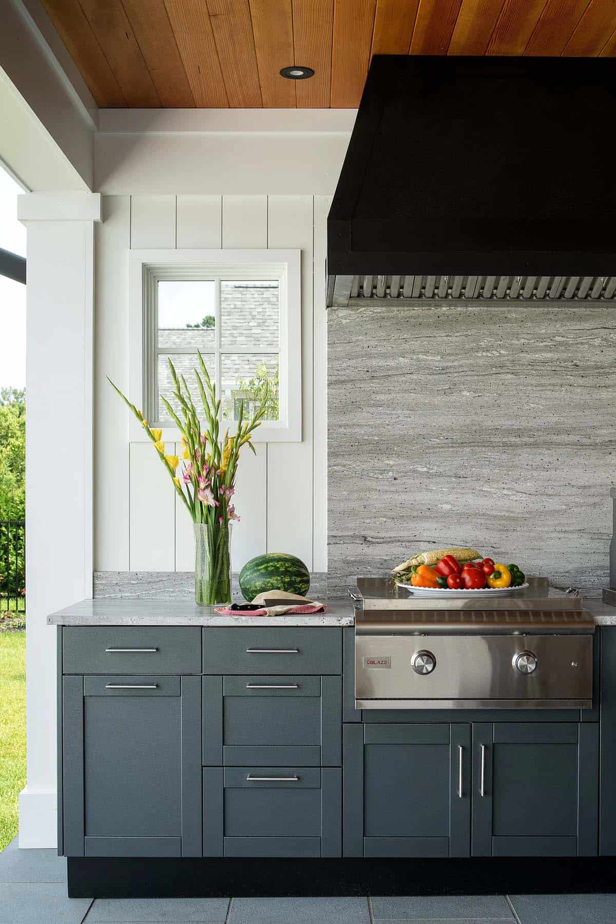 Outdoor kitchen with charcoal cabinets, built-in grill, quartzite backsplash, and black range hood under a cedar ceiling
