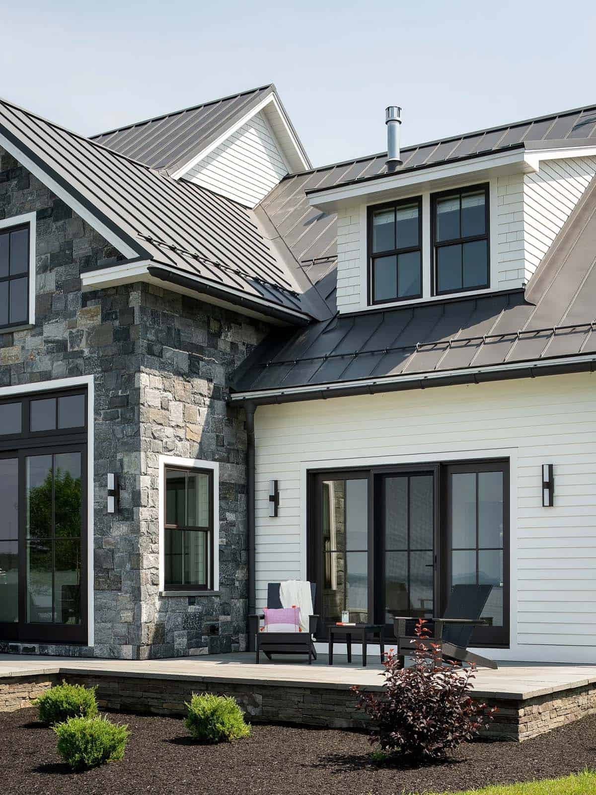 Stone and white clapboard exterior with black-framed windows, a shingled dormer, and Adirondack chairs on a stone patio