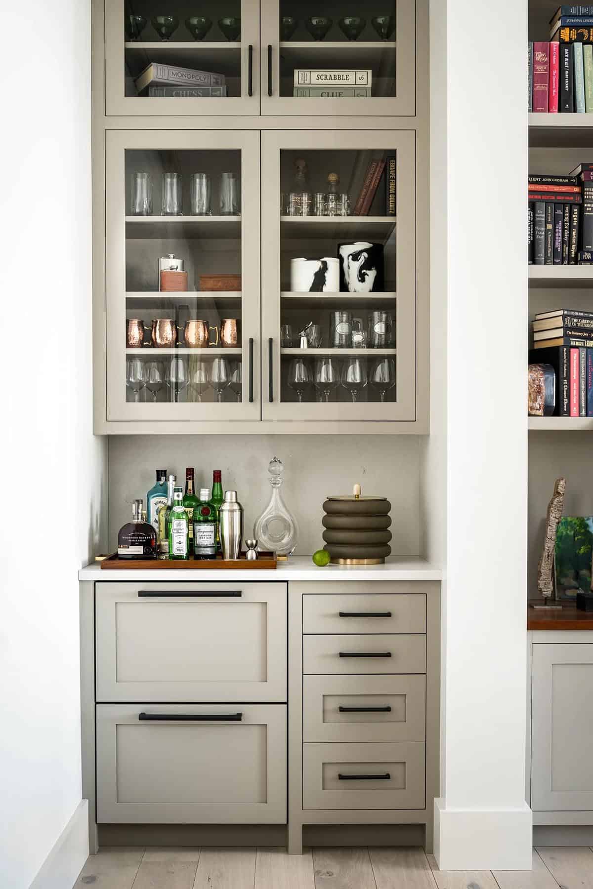 Built-in bar with glass-front greige cabinets, drink drawers, and adjacent bookshelves