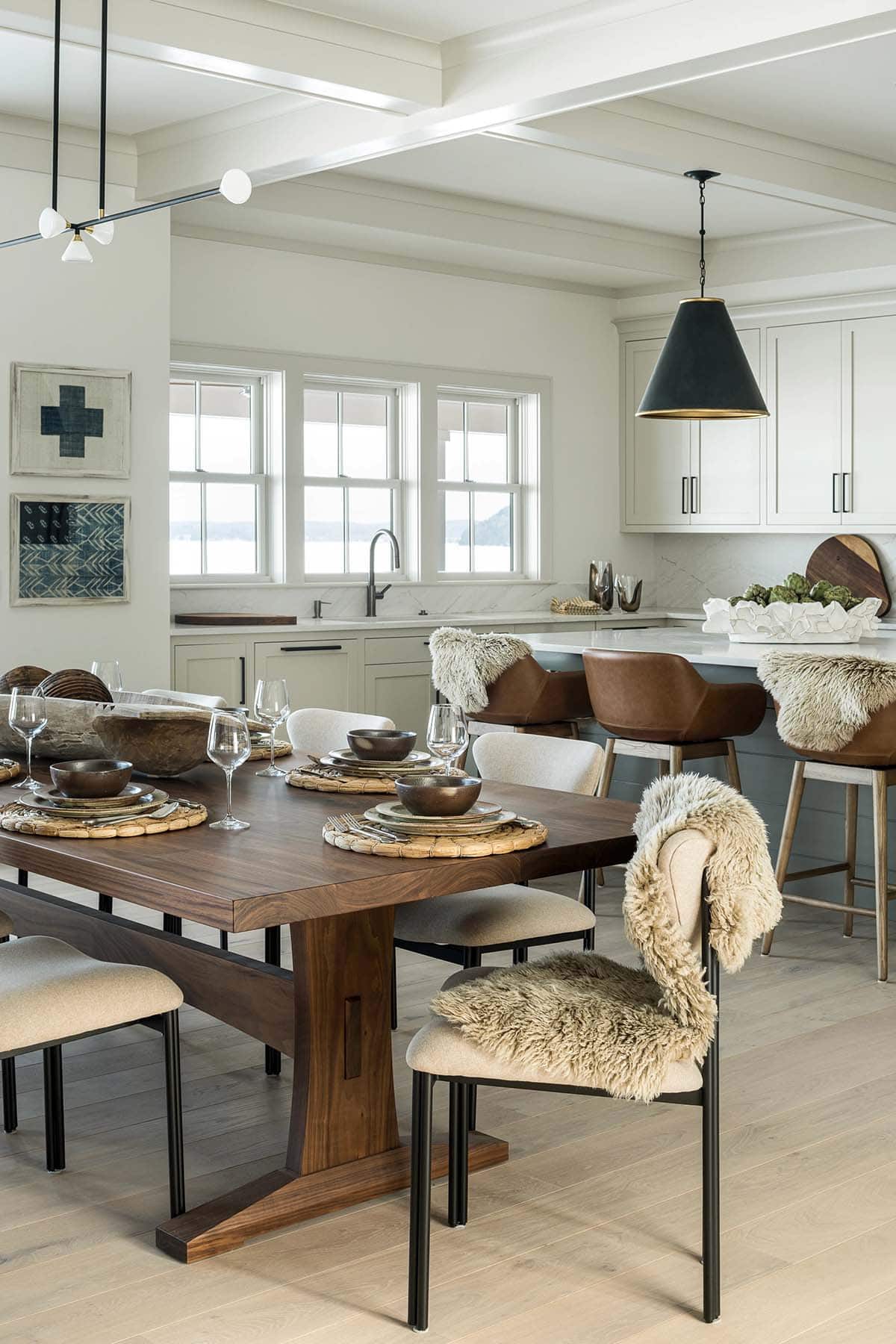 Walnut trestle dining table with upholstered chairs and sheepskins, open to the kitchen with lake views in winter