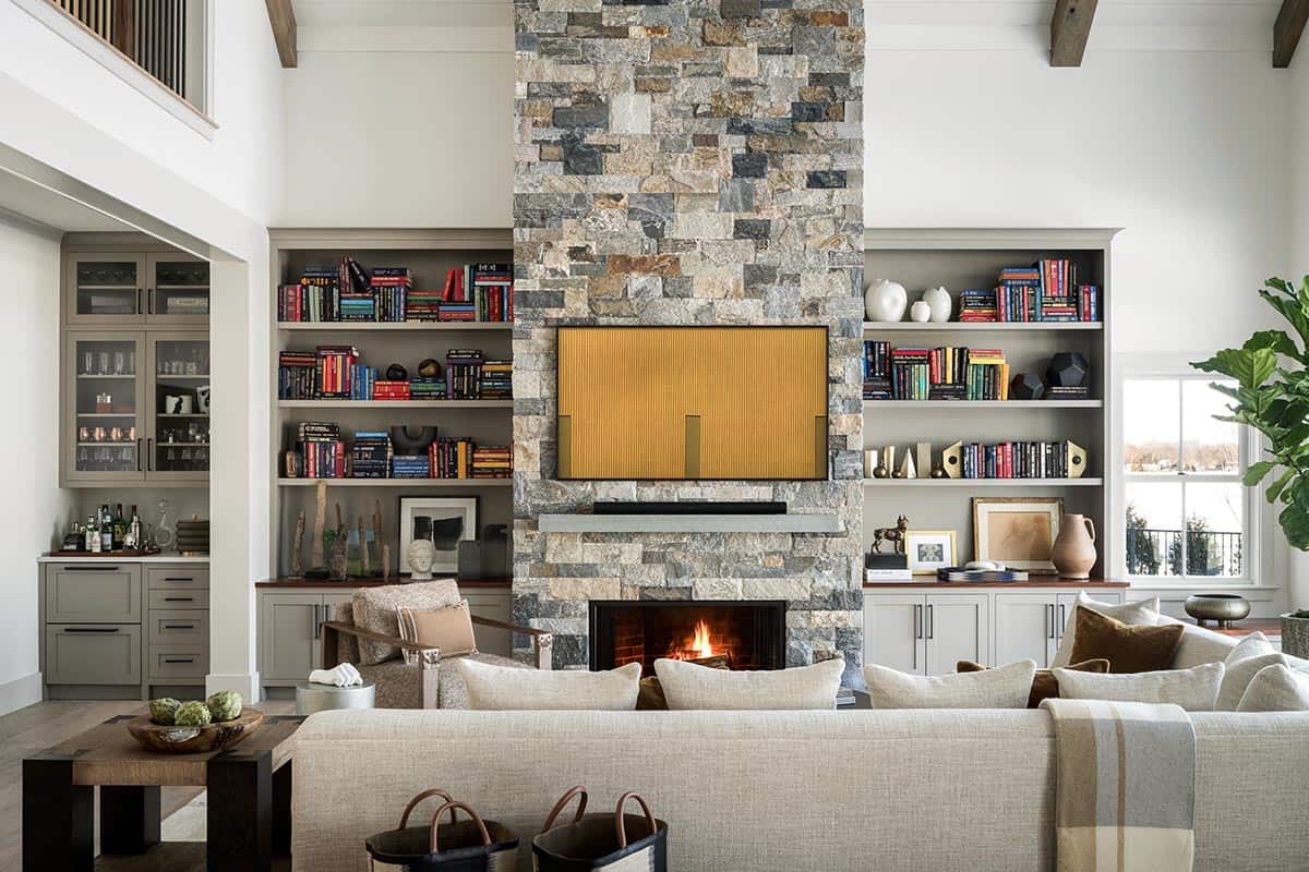 Double-height living room with stone fireplace, built-in bookshelves, wood ceiling beams, and a cream sectional sofa