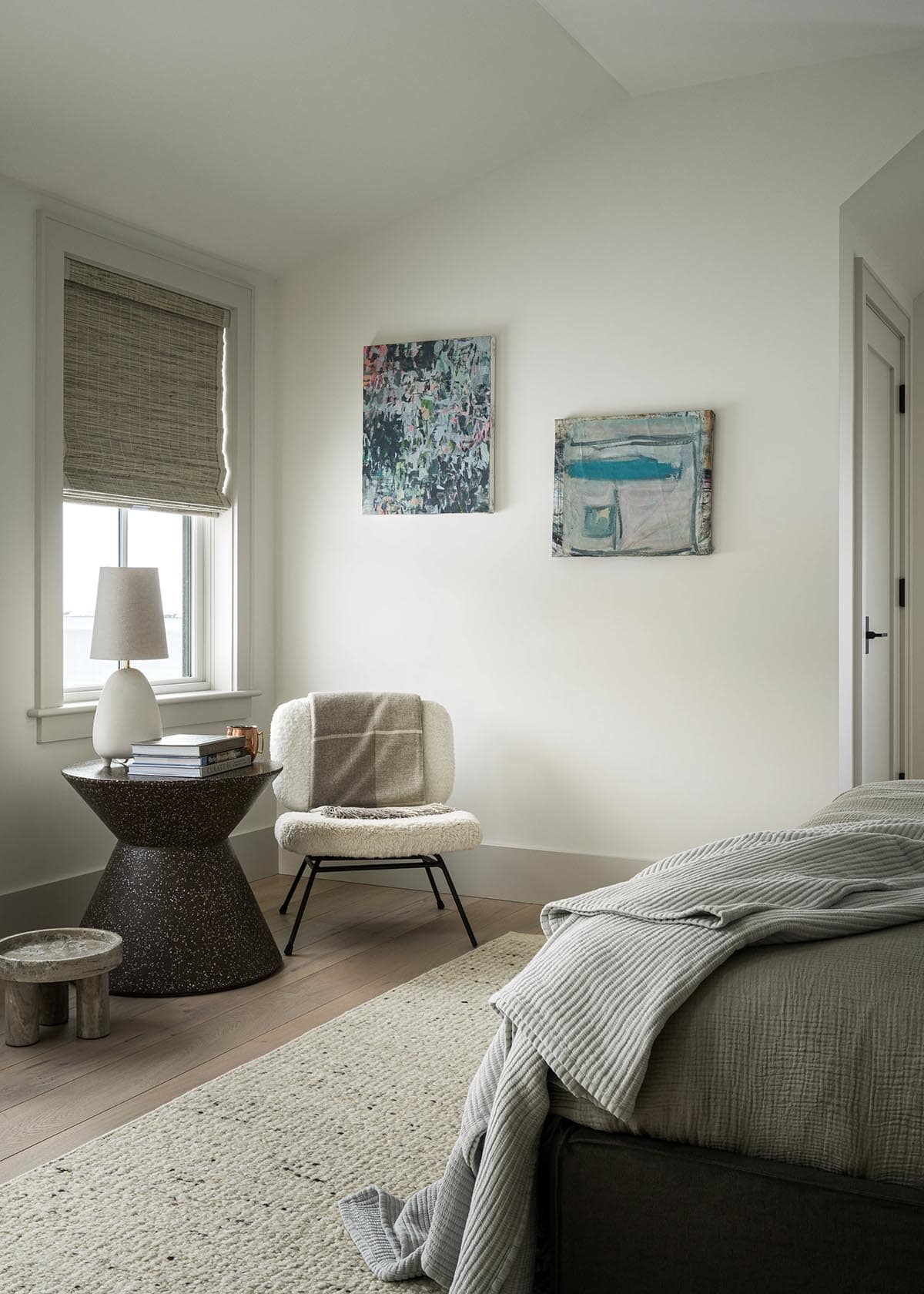 Guest bedroom corner with sherpa accent chair, speckled hourglass side table, abstract artwork, and woven shade