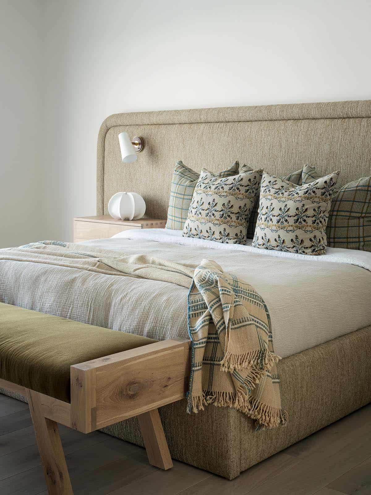 Bedroom with upholstered headboard, swing-arm sconce, patterned pillows, and an oak bench with olive velvet seat