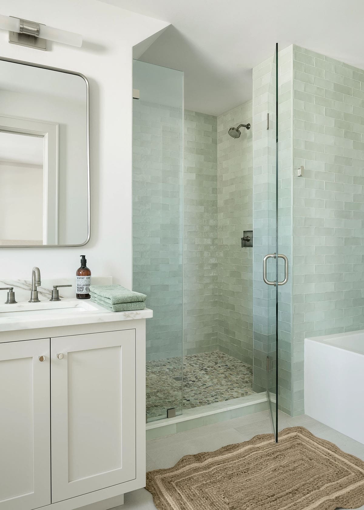 Bathroom with sage green tile walk-in shower, pebble floor, marble-topped white vanity, and jute rug