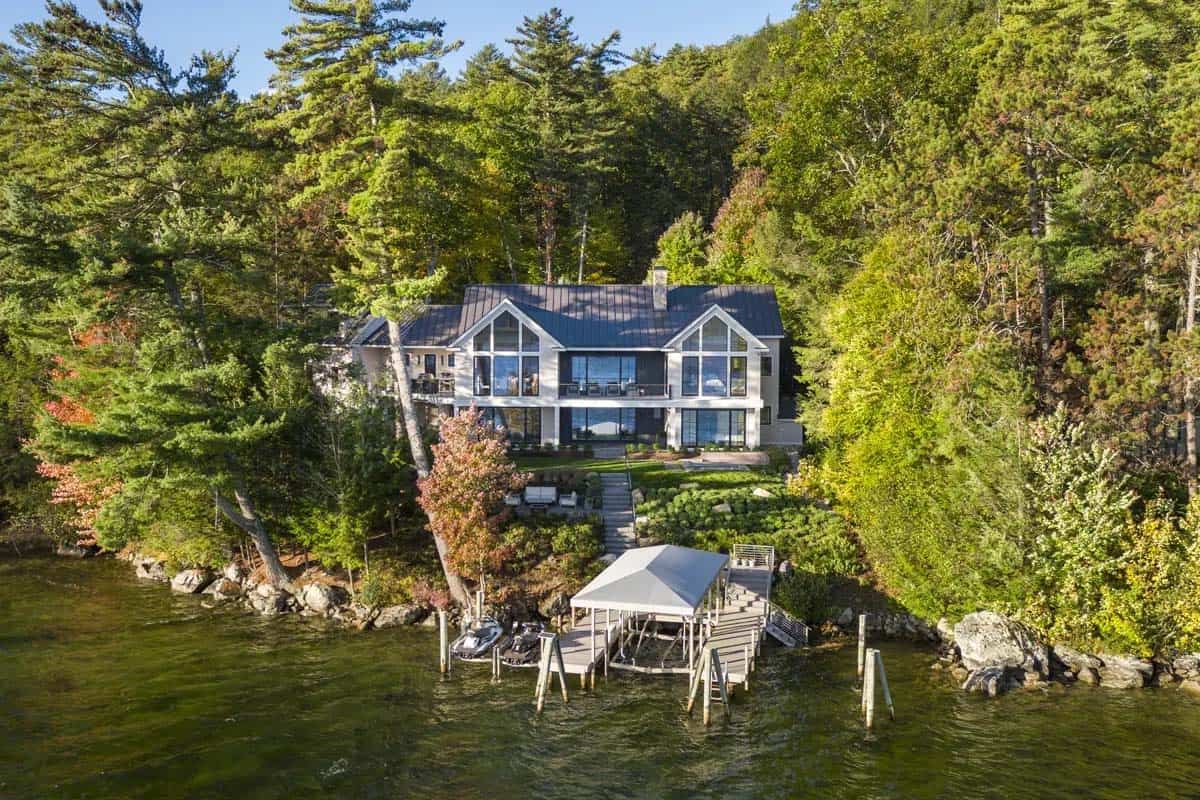 Aerial view of a modern New Hampshire lake house with a dock, boat lifts, and terraced landscaping surrounded by fall foliage