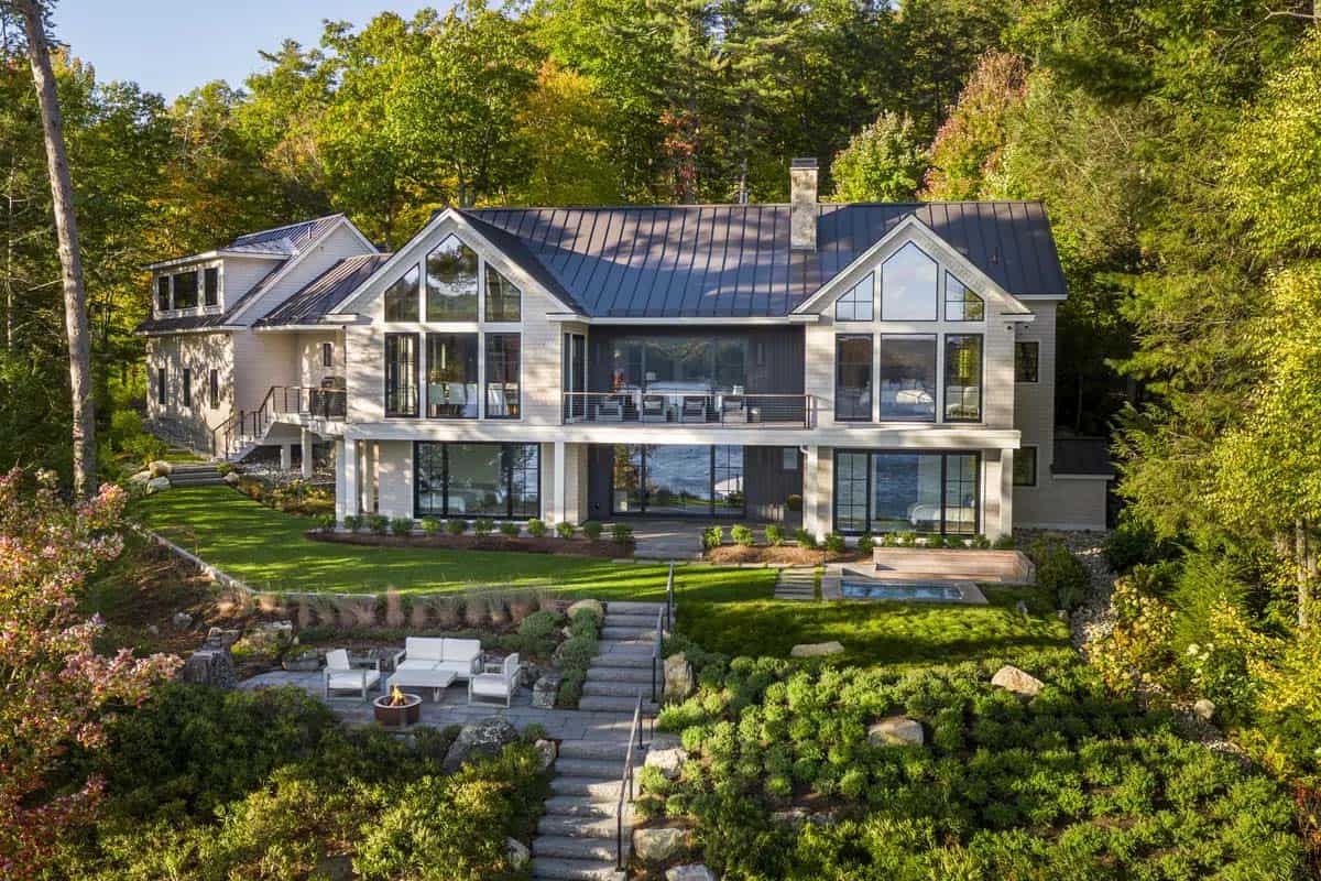 New Hampshire lake house rear facade with large gabled windows, metal roof, and stepped stone path down to a fire pit patio