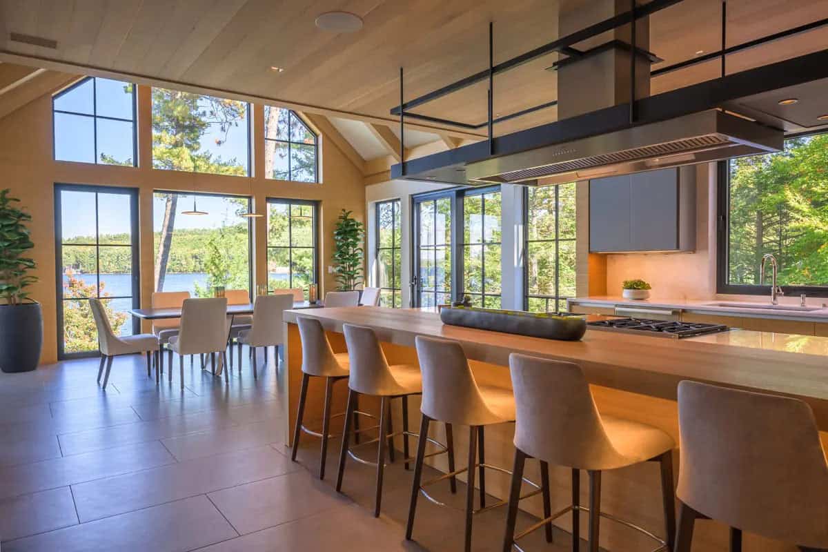 Kitchen island with bar stools and dining table beneath tall windows framing the lake and trees
