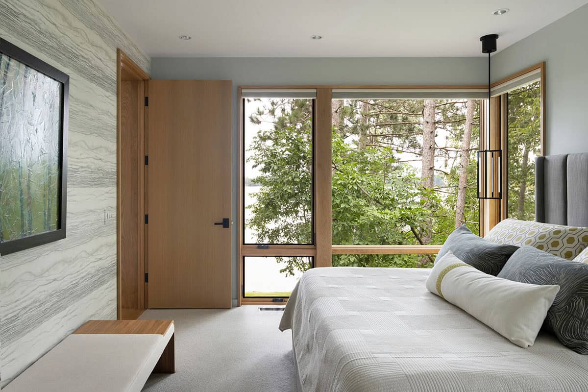Bedroom with wood-framed corner windows, upholstered headboard, and a tree-filled view