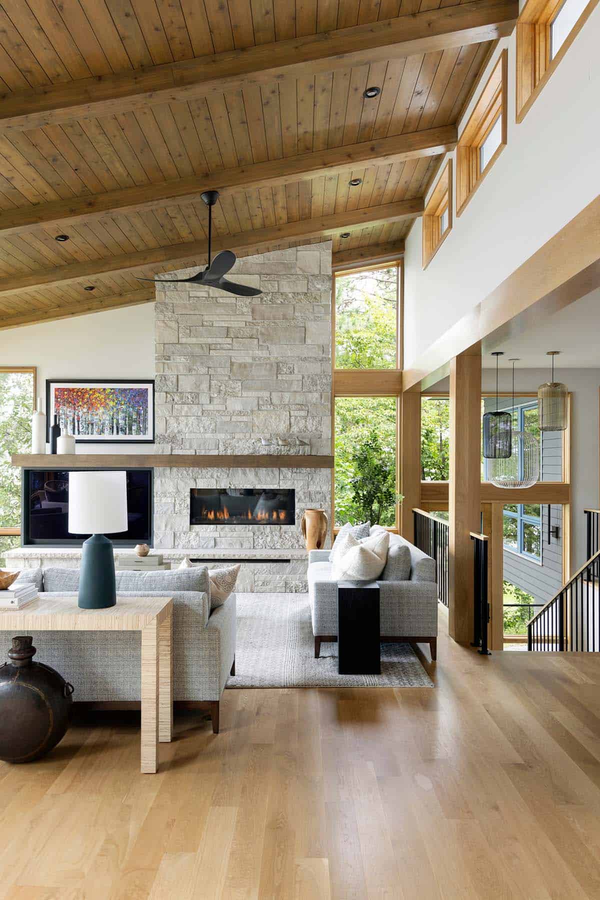 Stone fireplace with gas flames, ceiling fan, and tall windows in the vaulted living room