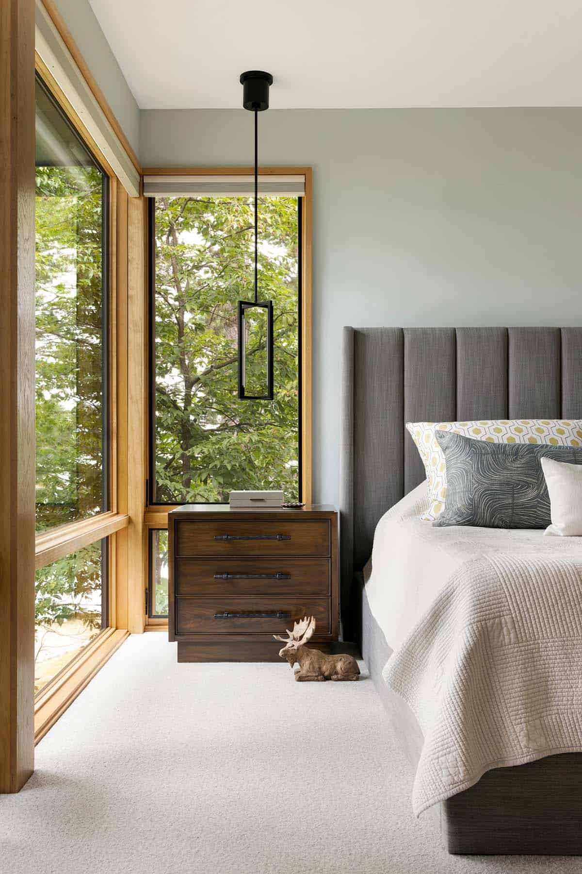 Primary bedroom corner with a tufted gray headboard, walnut nightstand, and tall tree-view windows
