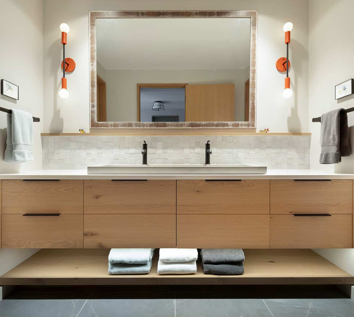 Double vanity with a floating oak cabinet, orange sconces, and a rustic framed mirror