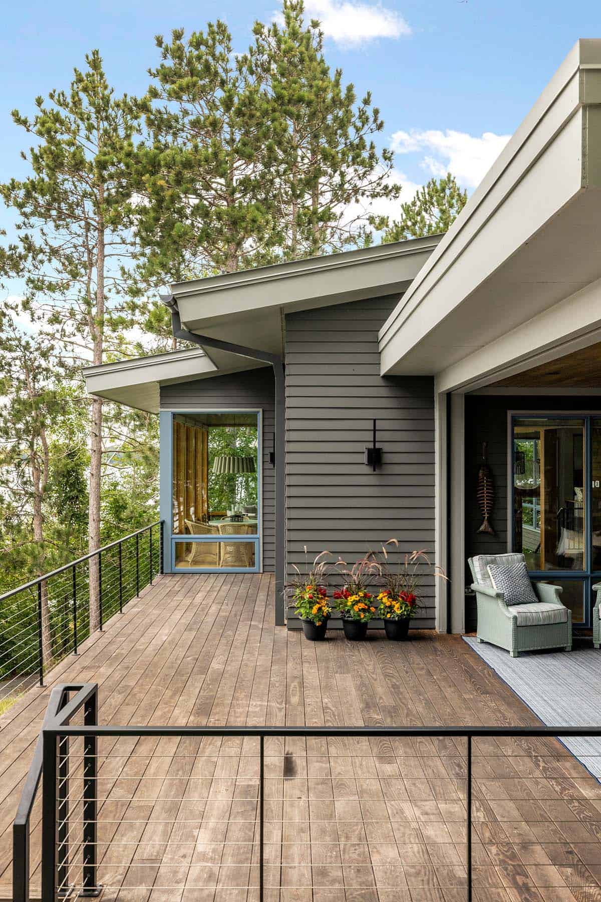 Deck corner with potted flowers, a sage wicker chair, and a blue-framed window beside the covered porch