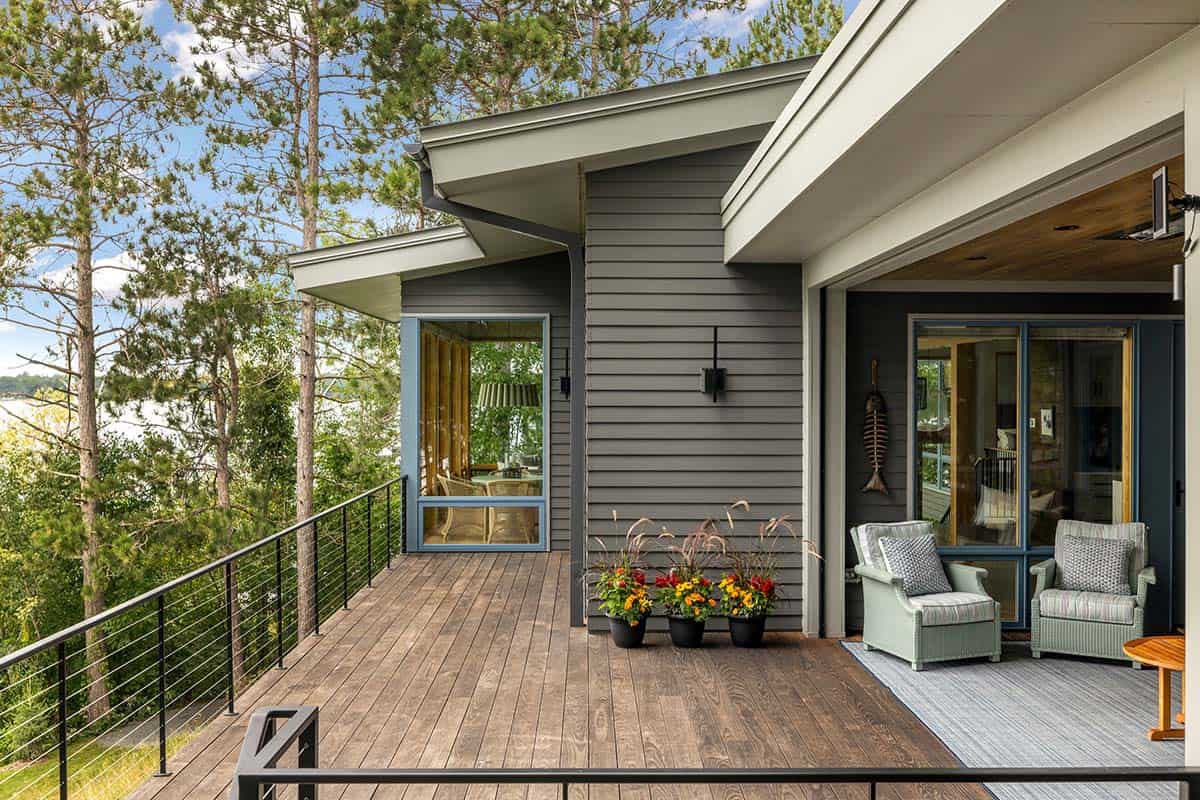 Gray-sided deck with flower planters and sage chairs under a roof overhang, with pines behind