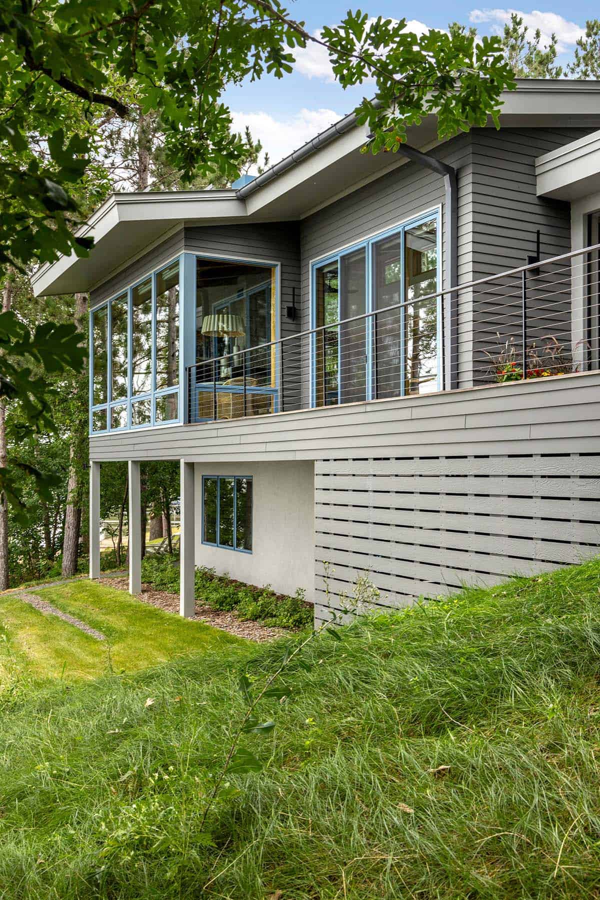 Side view of the house on its hillside, with blue-framed windows, deck posts, and a slatted wall