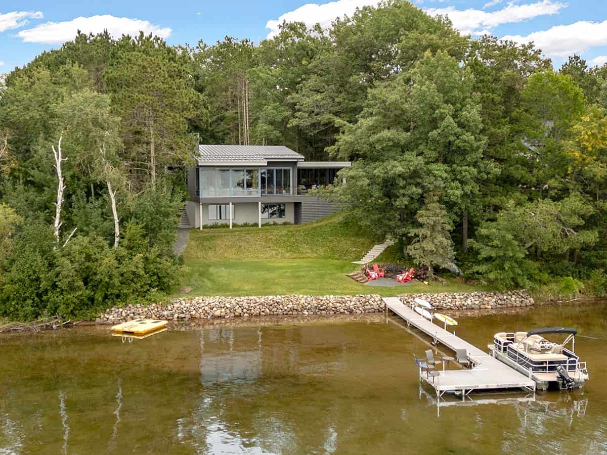 Aerial view of the lake house on its grassy slope, with a stone shoreline, dock, and pontoon boat
