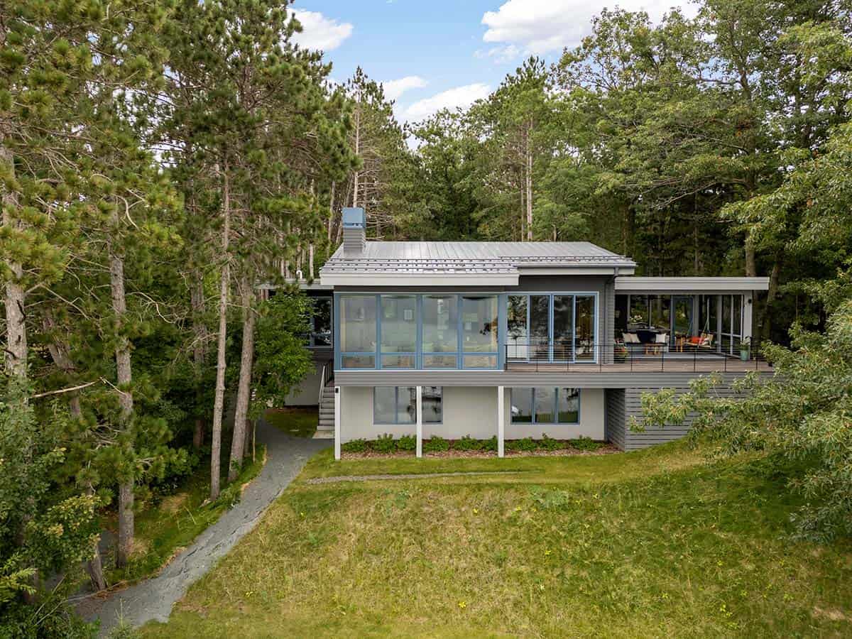 Gray lake house with blue window frames and a standing-seam metal roof, framed by tall pines