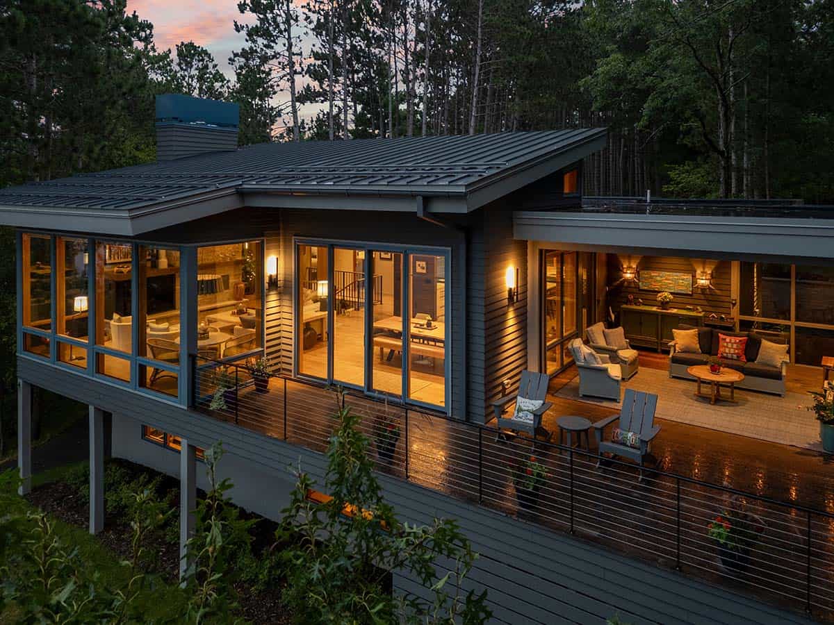 Lake house at dusk with a glowing great room, lit covered porch, and wet wood deck