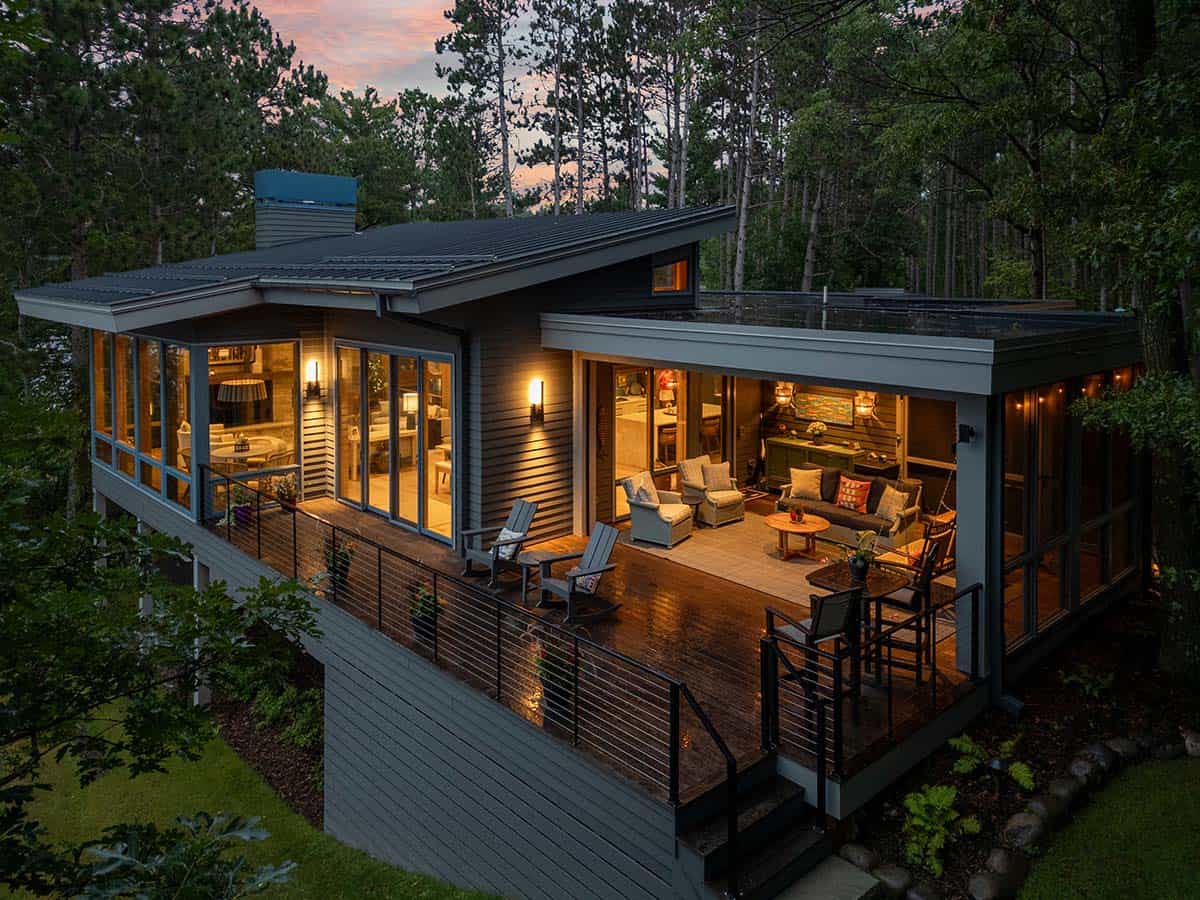 Aerial dusk view of the deck, lit living room, and porch seating among the pines