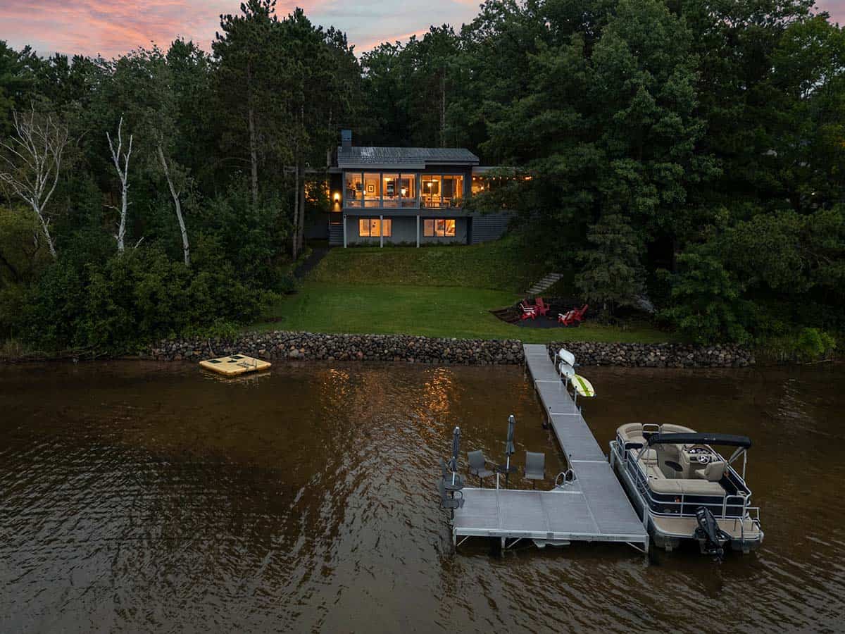Lake house glowing at twilight above the lawn, with a long dock and pontoon boat