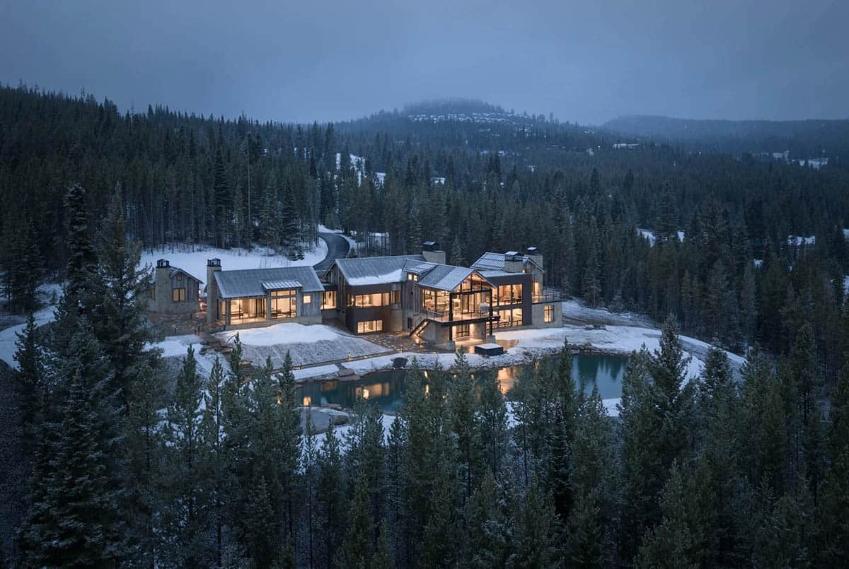 Aerial view of a mountain home beside a pond in a snowy fir forest at dusk