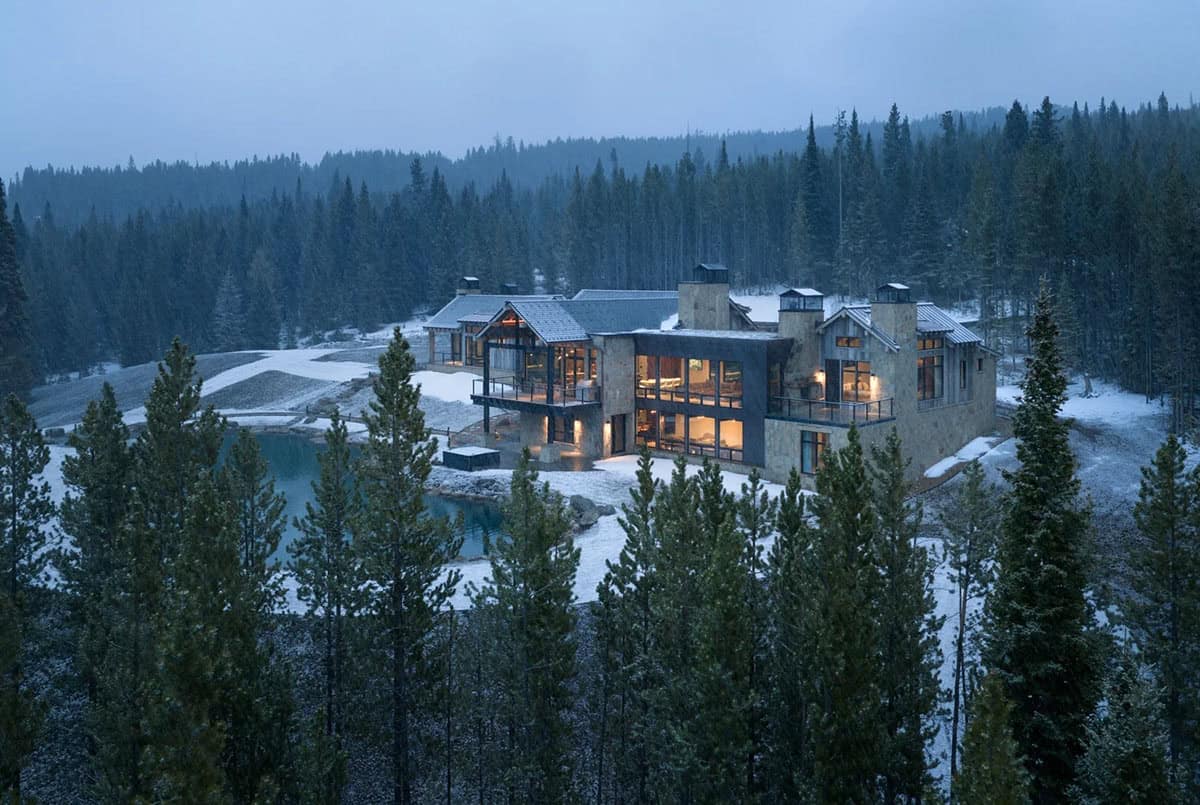 Mountain home glowing at dusk beside a pond in a snowy forest