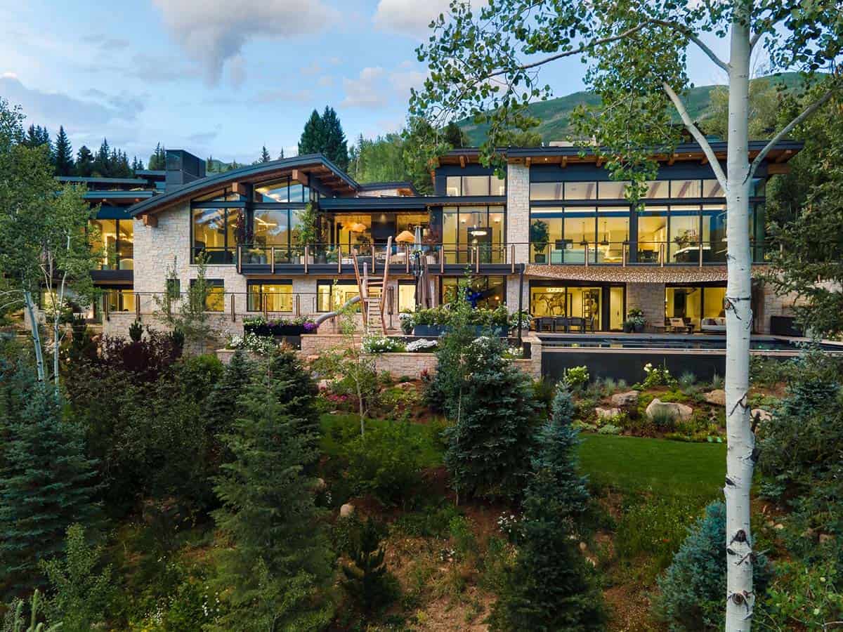 Mountain home with stone and glass facade, terraces, and pool glowing at dusk behind an aspen tree
