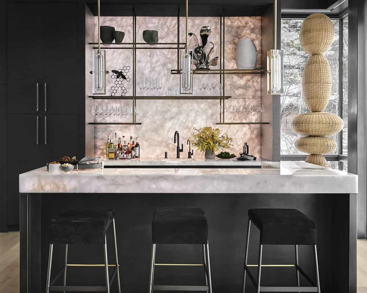 Home bar with a backlit onyx counter, brass shelving, black velvet stools, and a stacked wicker sculpture