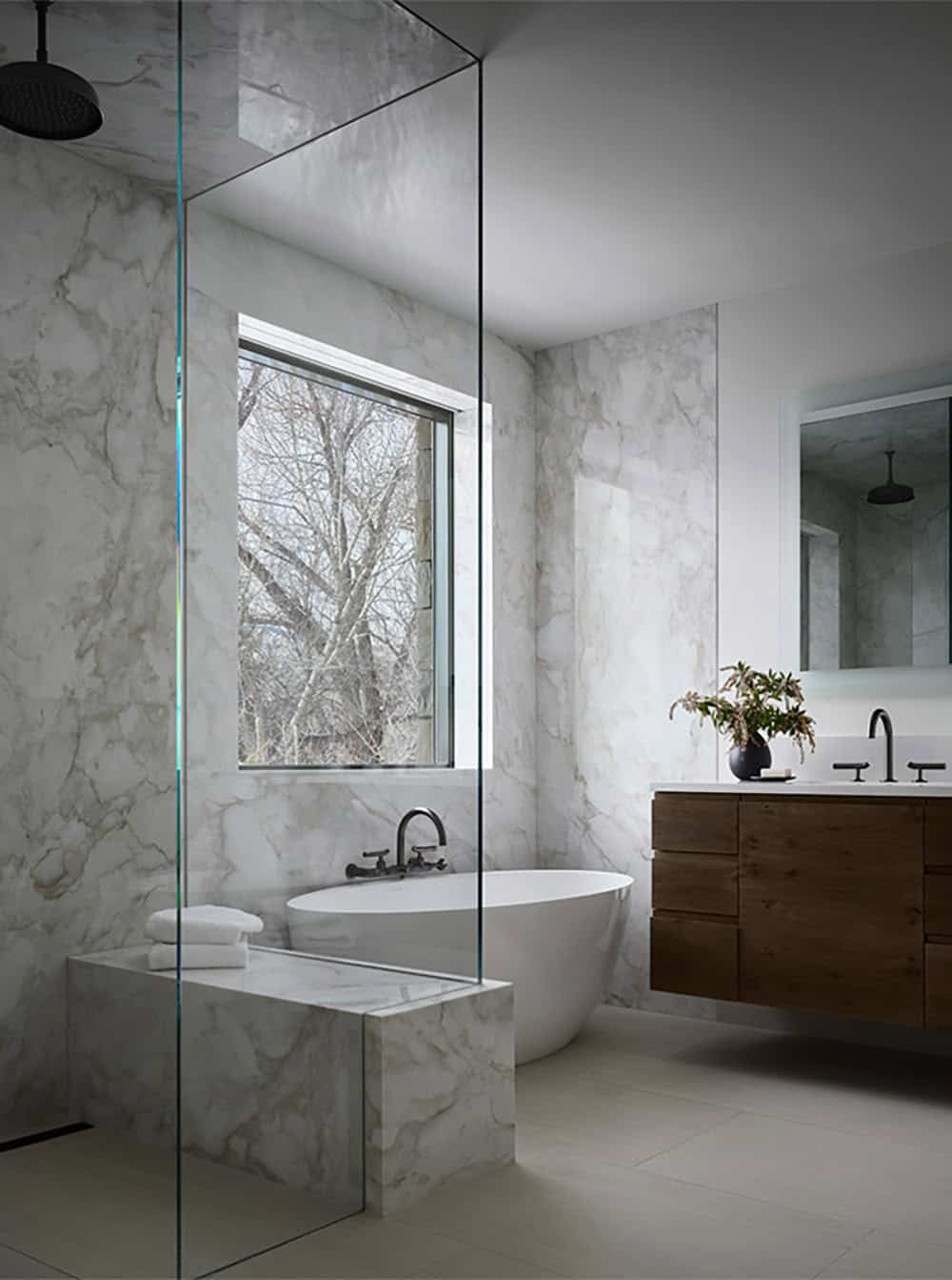 Marble-clad bathroom with a glass shower, freestanding tub, and floating wood vanity in winter light