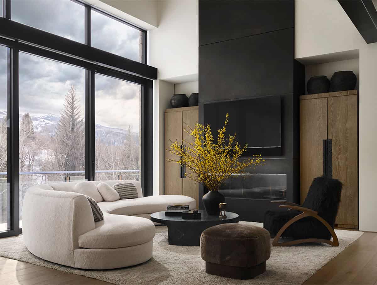Living room with a curved cream sofa, black fireplace wall, yellow forsythia branches, and snowy mountain views