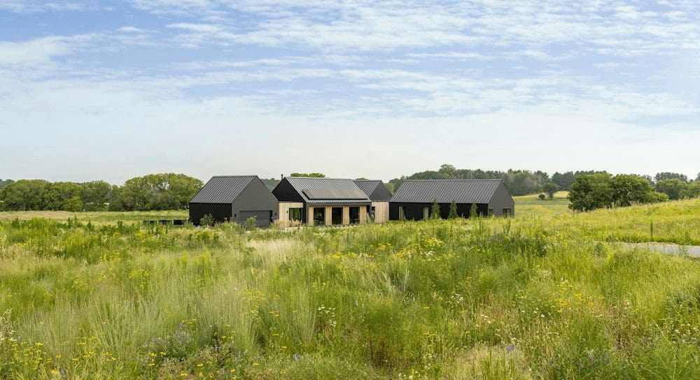 Distant view of the black and wood-clad prairie home nestled into green rolling grassland with wildflowers and a tree line