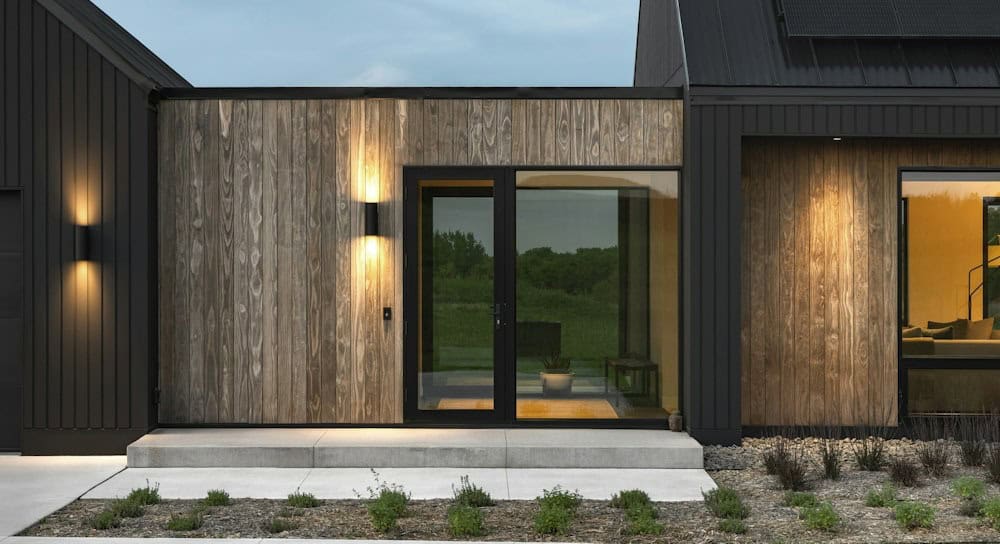 Front entry with weathered vertical wood siding, a black glass door and glowing cylinder wall sconces between black-sided gables