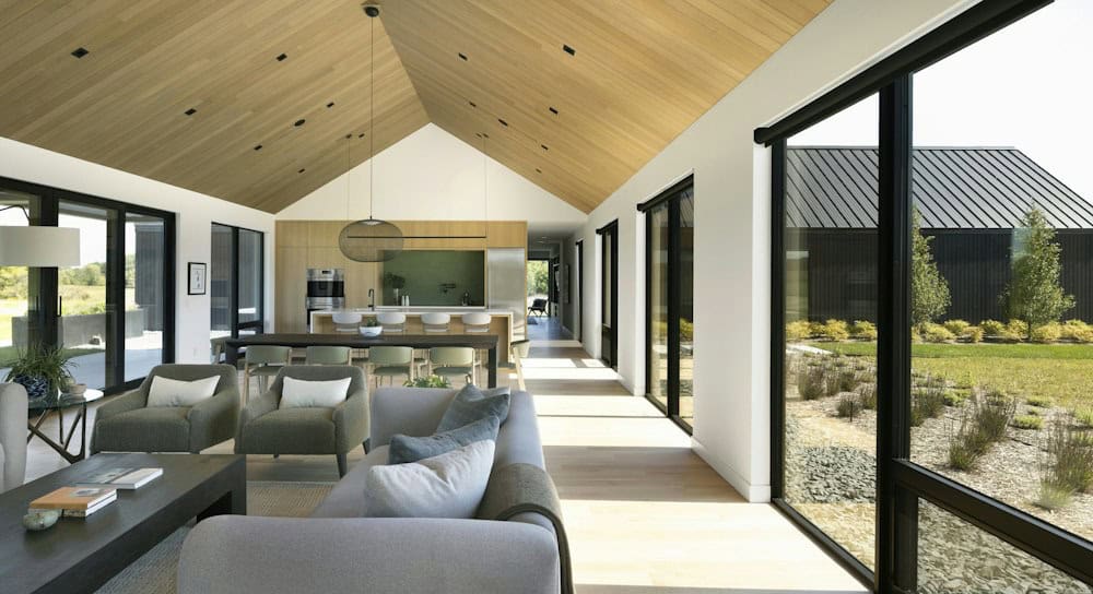 Long great room under a vaulted white oak ceiling with a gray sofa, olive armchairs, a dining table and kitchen beyond, with black-framed glass to the courtyard
