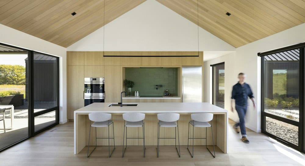White oak kitchen with a white waterfall island, four gray counter stools, a linear black pendant and a green tile niche under a vaulted wood ceiling