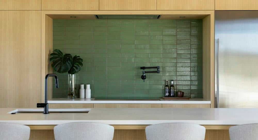 Glossy green stacked tile backsplash in a white oak niche with a black pot filler, monstera leaves in a vase and a black faucet on the island