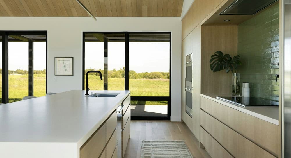 Kitchen island with a black faucet and flat-front white oak drawers beside an induction cooktop, with a black-framed window looking to the meadow