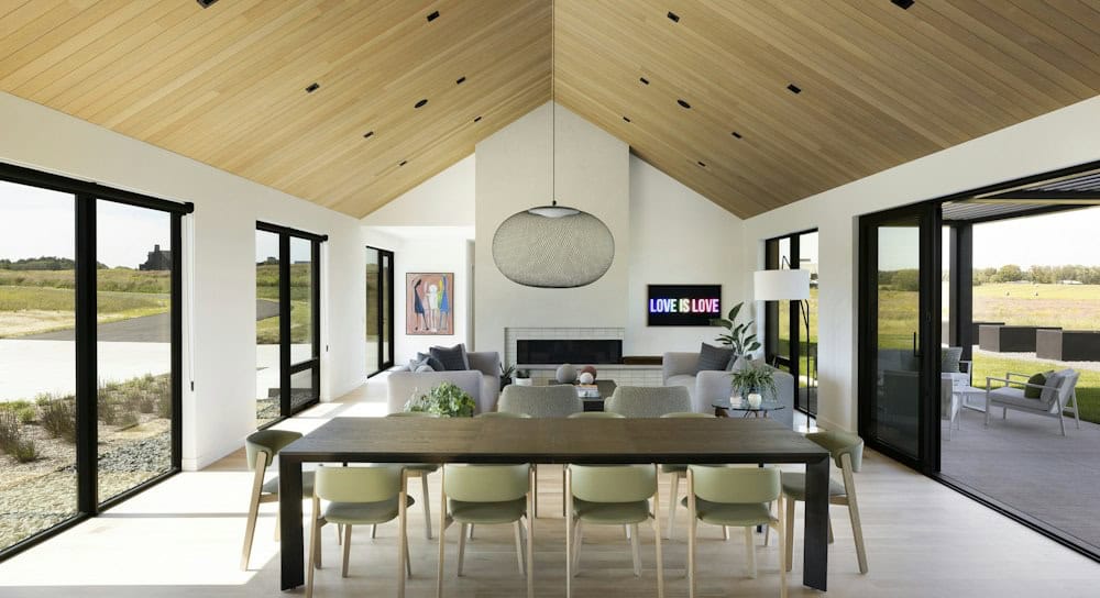 Dining room with a dark wood table and sage green chairs under a sculptural wire pendant, opening to a living area with a fireplace and neon art