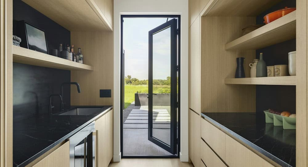Back kitchen and bar with white oak open shelves, black stone countertops and a glass door opening to a garden path and raised planters