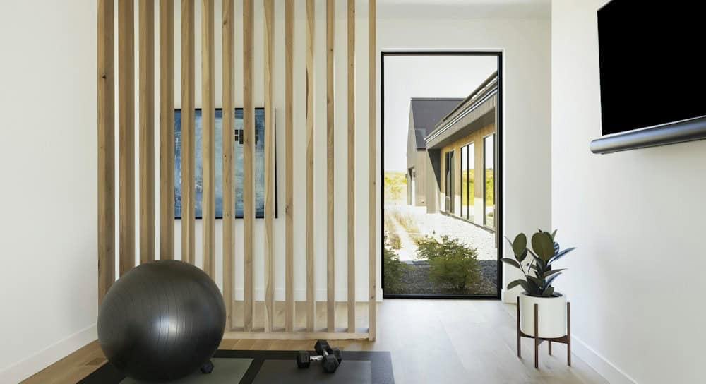 Exercise room with a white oak slat screen, a black exercise ball and dumbbells, and a tall window looking across the courtyard to another pavilion