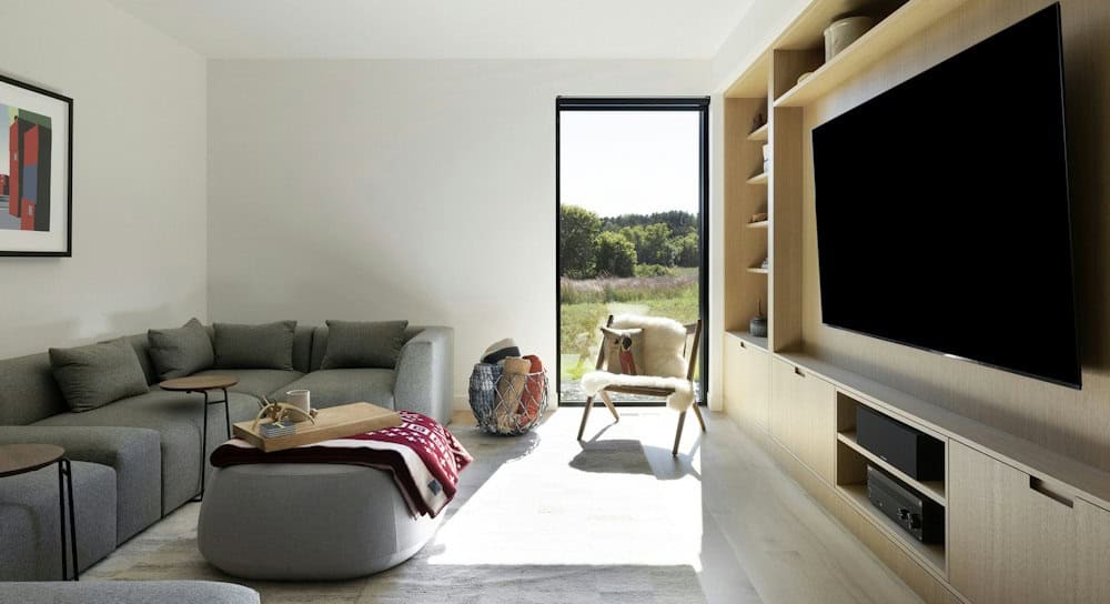 Den with a gray sectional and ottoman facing a white oak built-in media wall, with a sheepskin chair by a tall window to the meadow